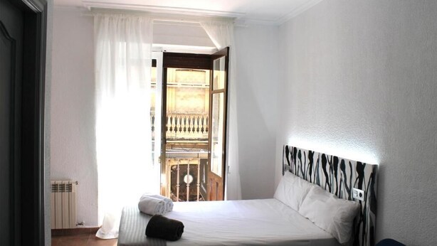 A bedroom with a bed, a radiator, a window with curtains, and a view of an outdoor balcony. - Alevia (Salamanca)