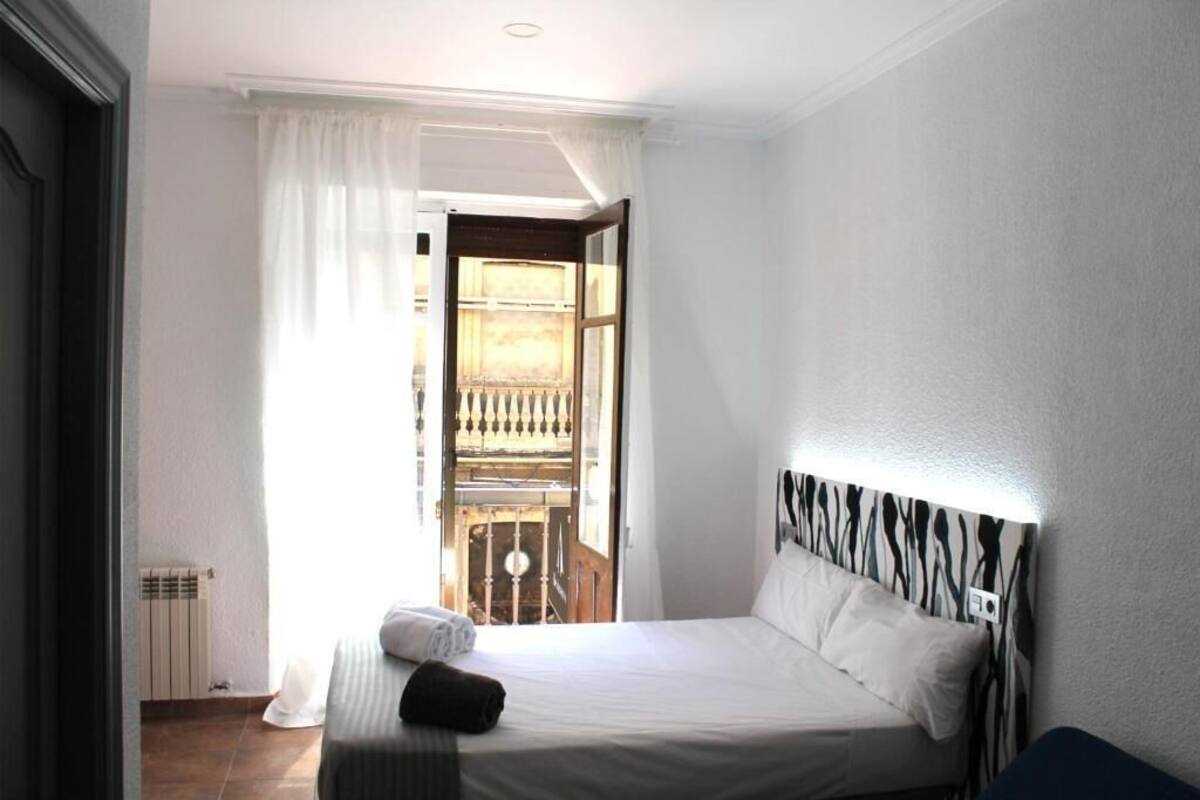 A bedroom with a bed, a radiator, a window with curtains, and a view of an outdoor balcony.