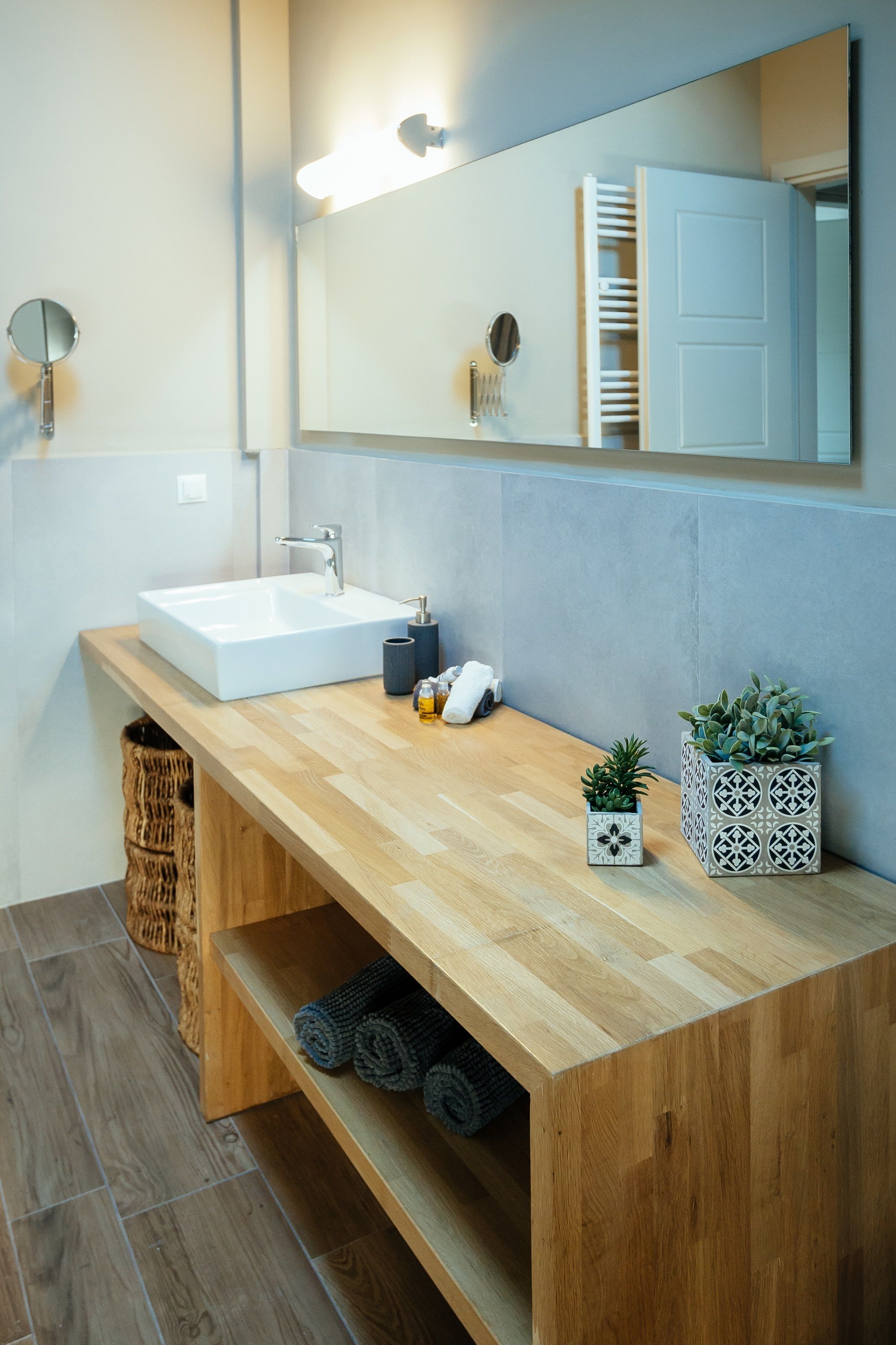 A modern bathroom with a wooden vanity, a white sink, and a mirror above it. There are two potted plants on the vanity, a towel rack, and a basket under the sink.