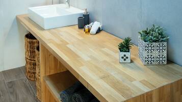 A modern bathroom with a wooden vanity, a white sink, and a mirror above it. There are two potted plants on the vanity, a towel rack, and a basket under the sink.