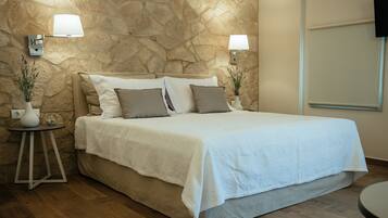 A neatly made bed with a white bedspread and two pillows, a stone wall, two wall-mounted lamps, a small round table with a vase, and a flat-screen TV mounted on the wall.