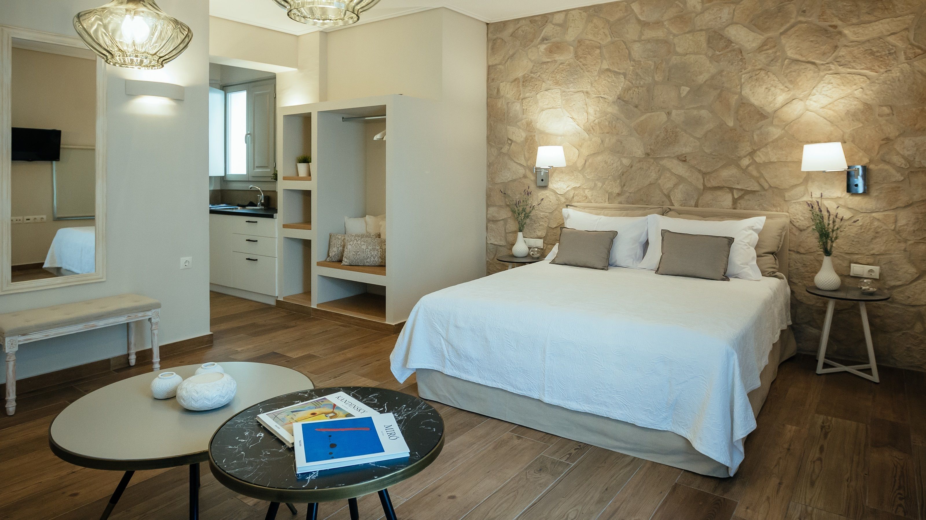 Een slaapkamer met een groot bed, een nachtkastje met boeken, een klein rond tafeltje, een spiegel, een kledingkast en een stenen muur.