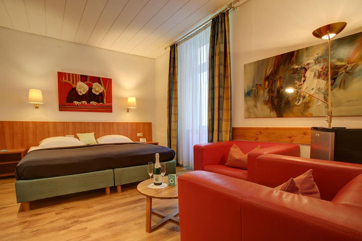 A hotel room with a bed, a red sofa, a small table with two bottles, and a painting on the wall.