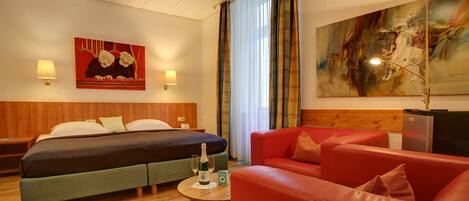 A hotel room with a bed, a red sofa, a small table with two bottles, and a painting on the wall.
