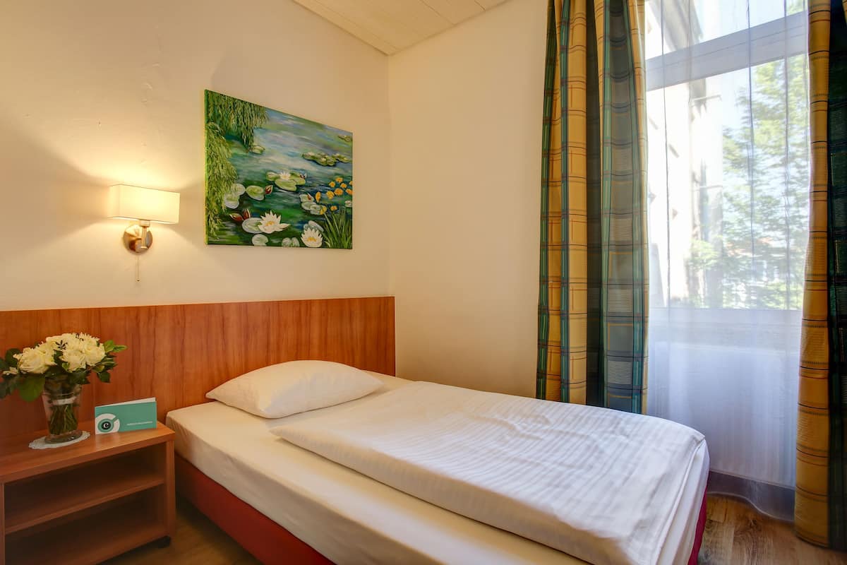 A hotel room with a bed, a nightstand with a vase of flowers, a painting of water lilies, and a window with curtains.