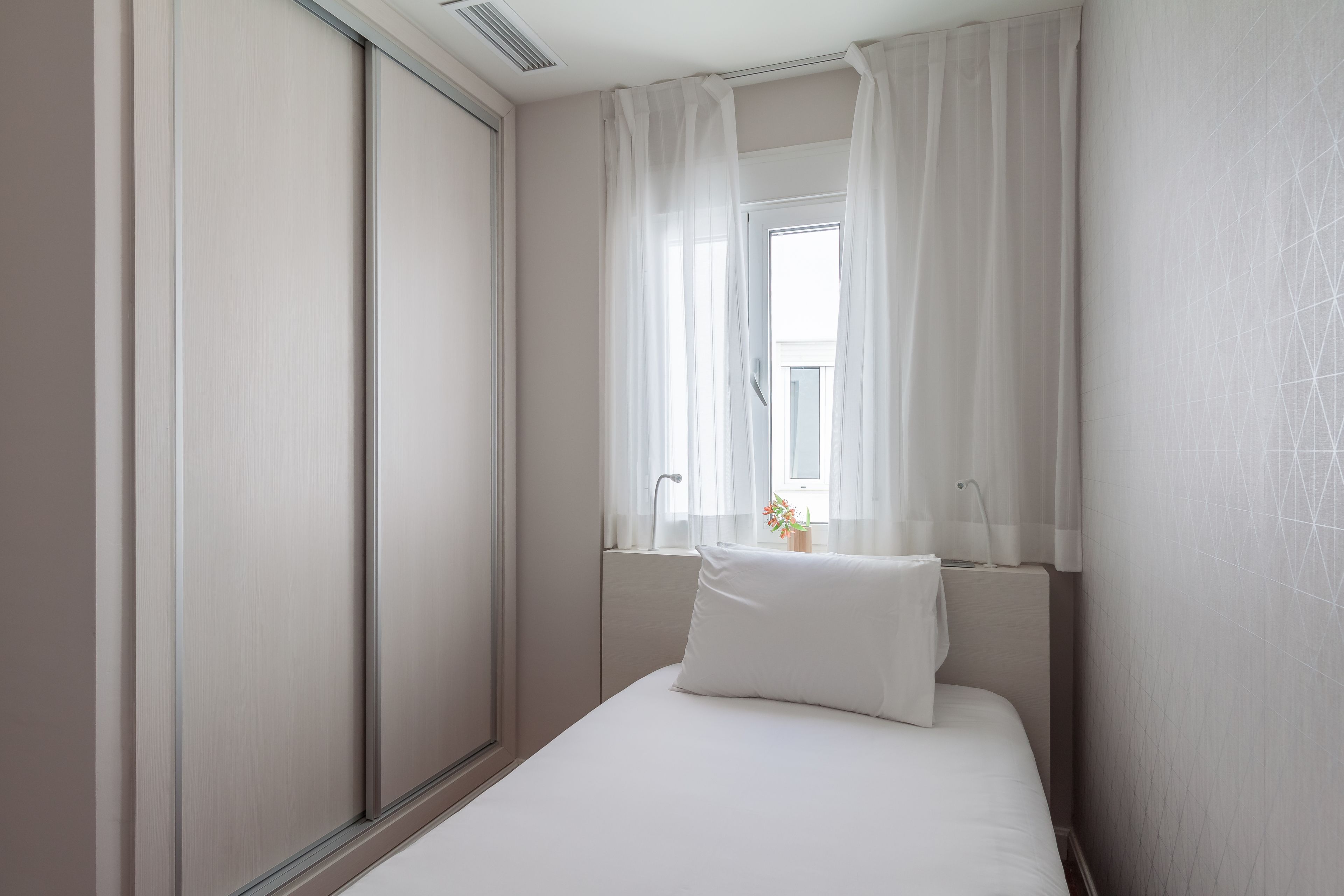Un dormitorio con cama, una ventana con cortinas traslúcidas, una mesita de noche y un radiador fijado a la pared.