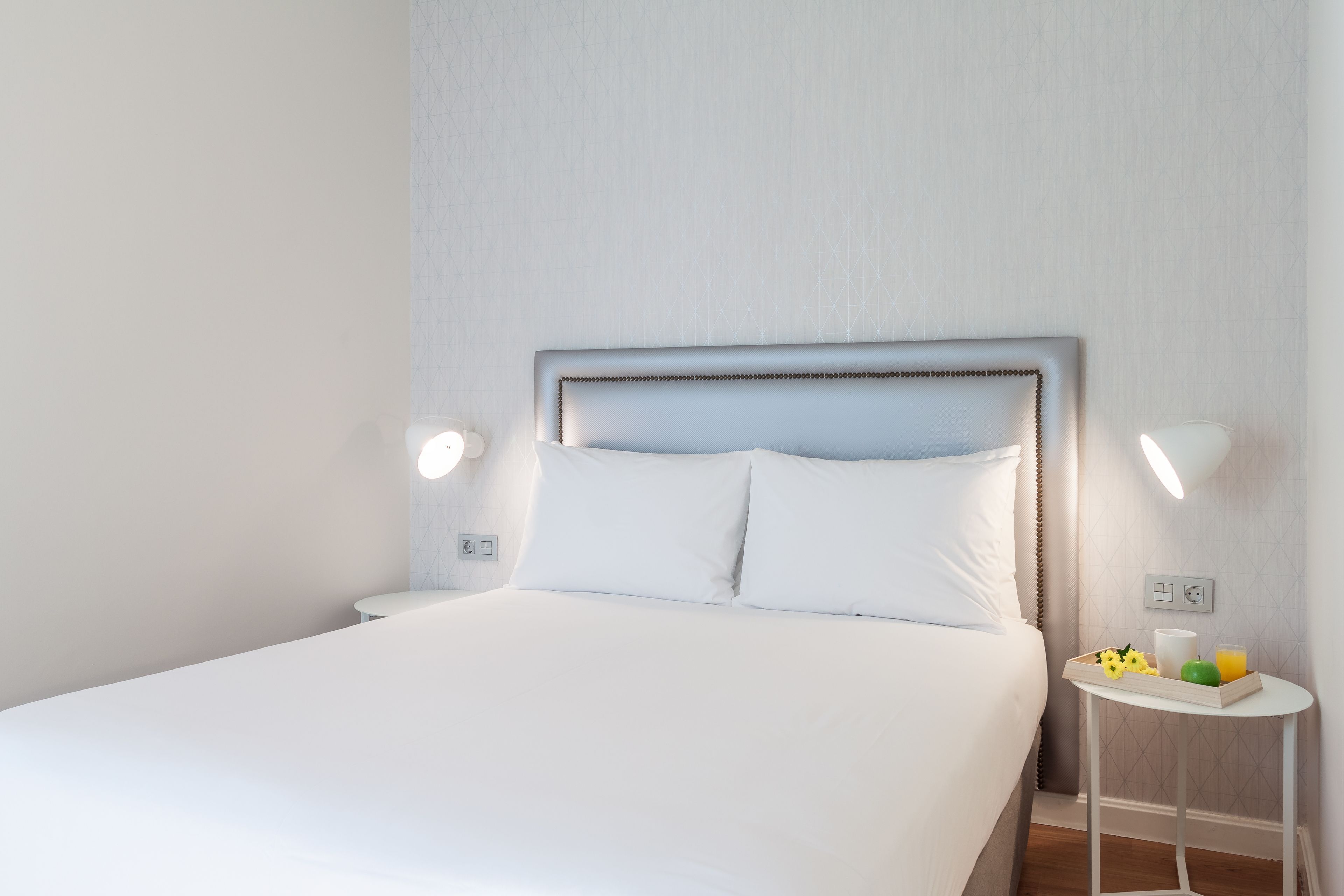 Una cama bien hecha con sábanas blancas, una mesita de noche con una bandeja de desayuno y lámparas fijadas a la pared.