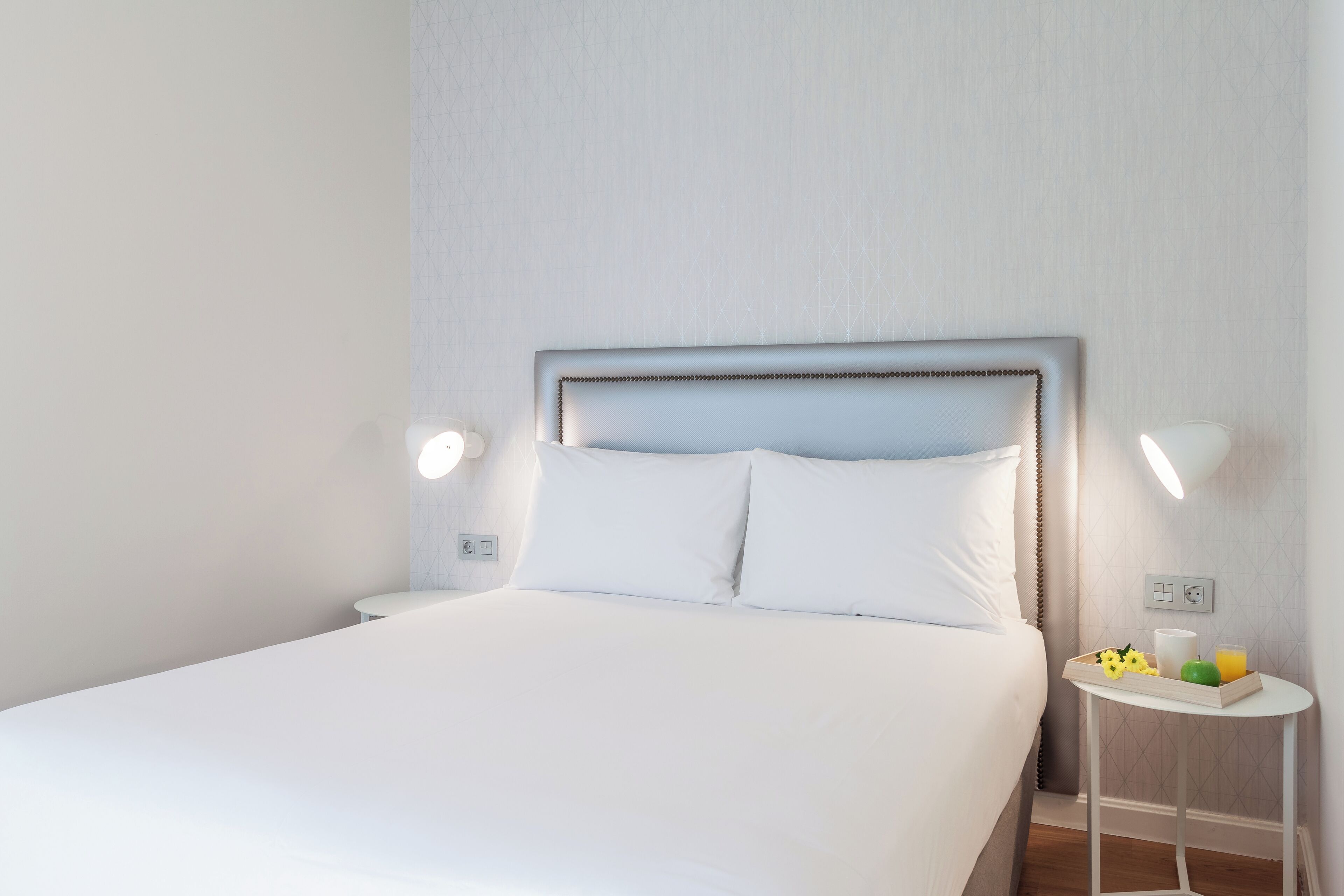 A neatly made bed with white linens, a bedside table with a tray of breakfast items, and two wall-mounted lamps.