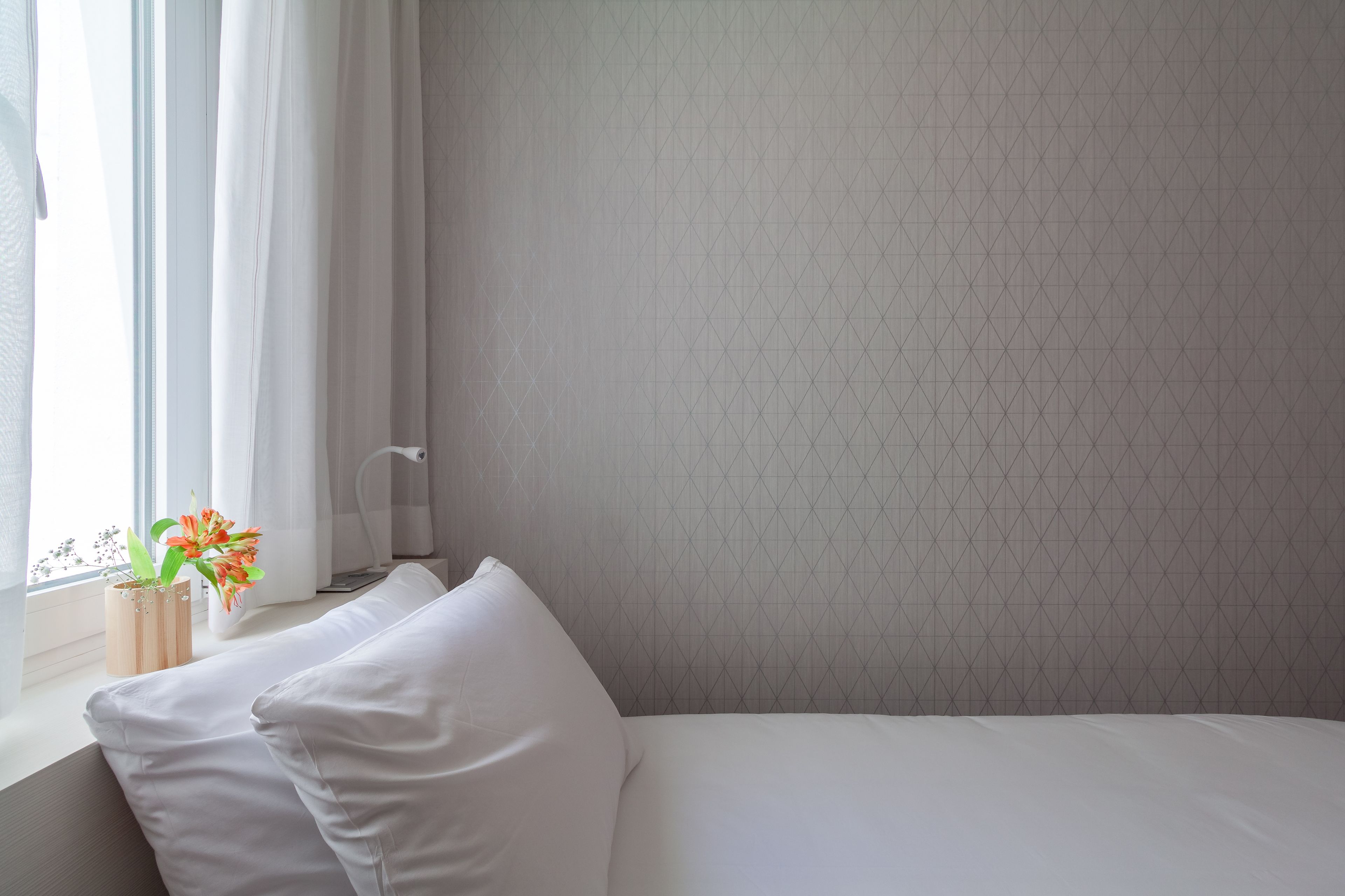 Una cama bien hecha con sábanas blancas, una ventana con cortinas traslúcidas y una mesita de madera con un jarrón de flores.