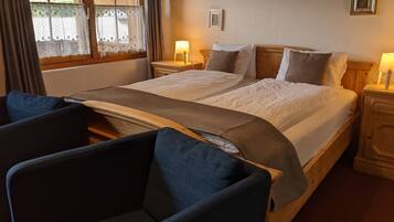 A double bed with a wooden headboard, two bedside tables with lamps, a chair, and a window with lace curtains.