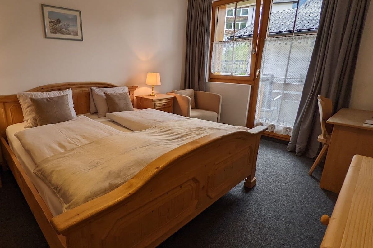 A bedroom with a wooden bed, two bedside tables with lamps, a chair, and a window with curtains.