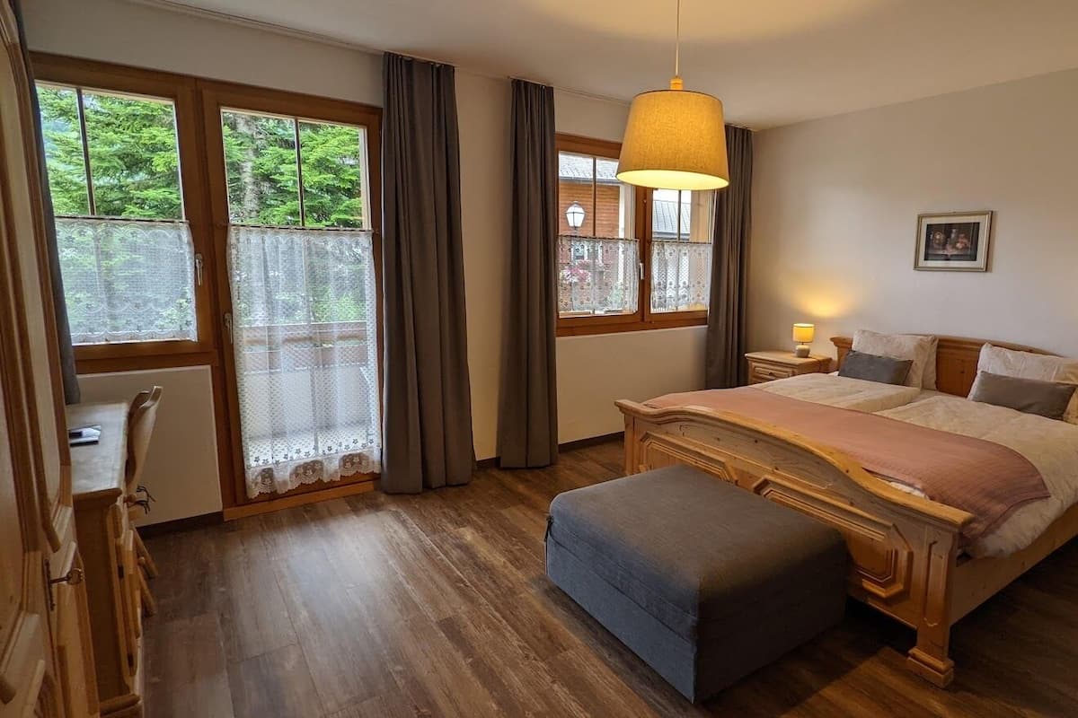 A bedroom with a wooden bed, a bench, a nightstand, a lamp, and a window with curtains.