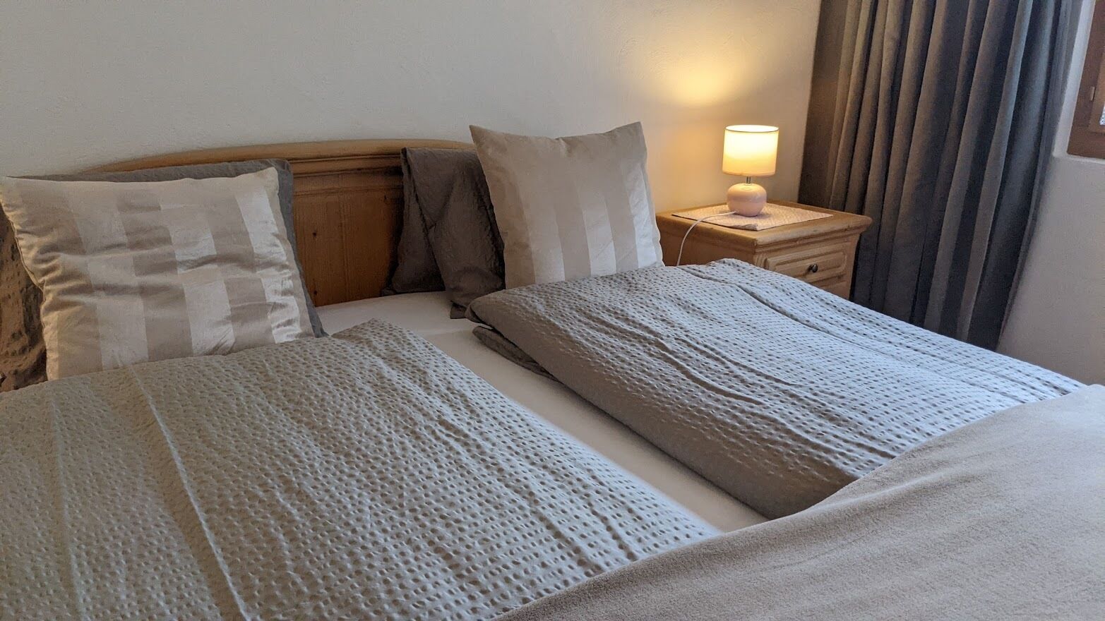 Una cama bien tendida con una manta texturizada y almohadas, una mesita de noche con lámpara, y una ventana con cortinas.