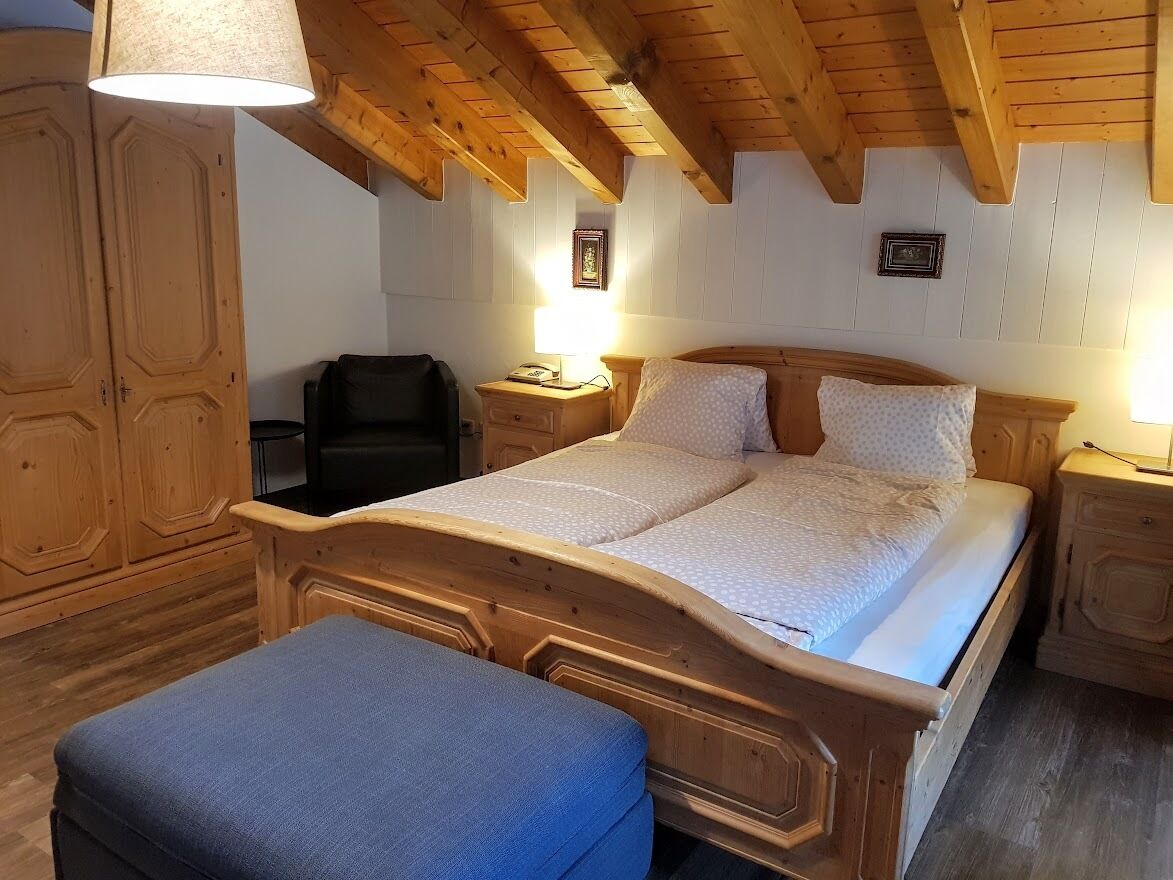 A wooden bed with a headboard, a bedside table, a lamp, and a wardrobe in a room with wooden beams on the ceiling.
