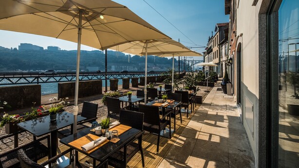 An outdoor terrace with tables and chairs, large umbrellas, and a view of a river and city skyline. - Vila Gale Porto Ribeira Hotel (Porto)
