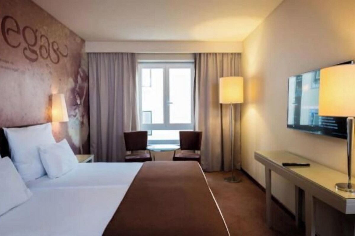 A hotel room with a large bed, a desk, a chair, a TV, and a window with curtains.