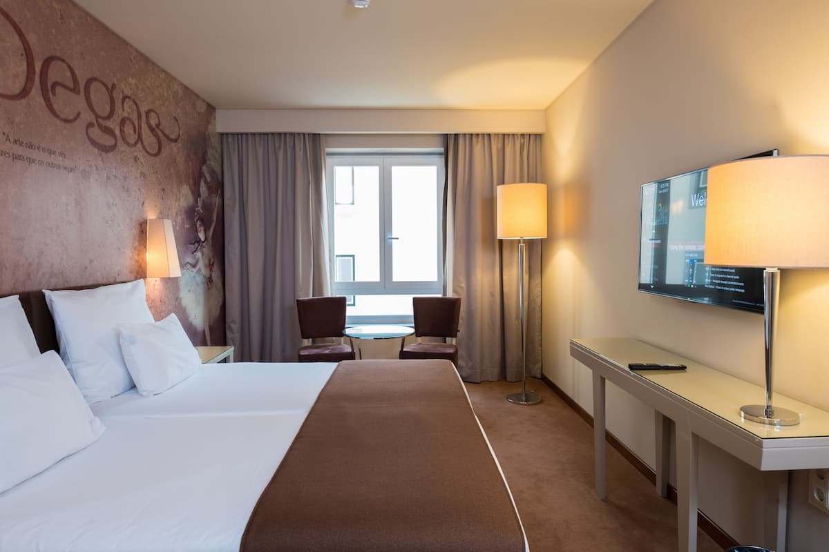 A hotel room with a large bed, two chairs, a desk with a lamp, a TV, and a window with curtains.