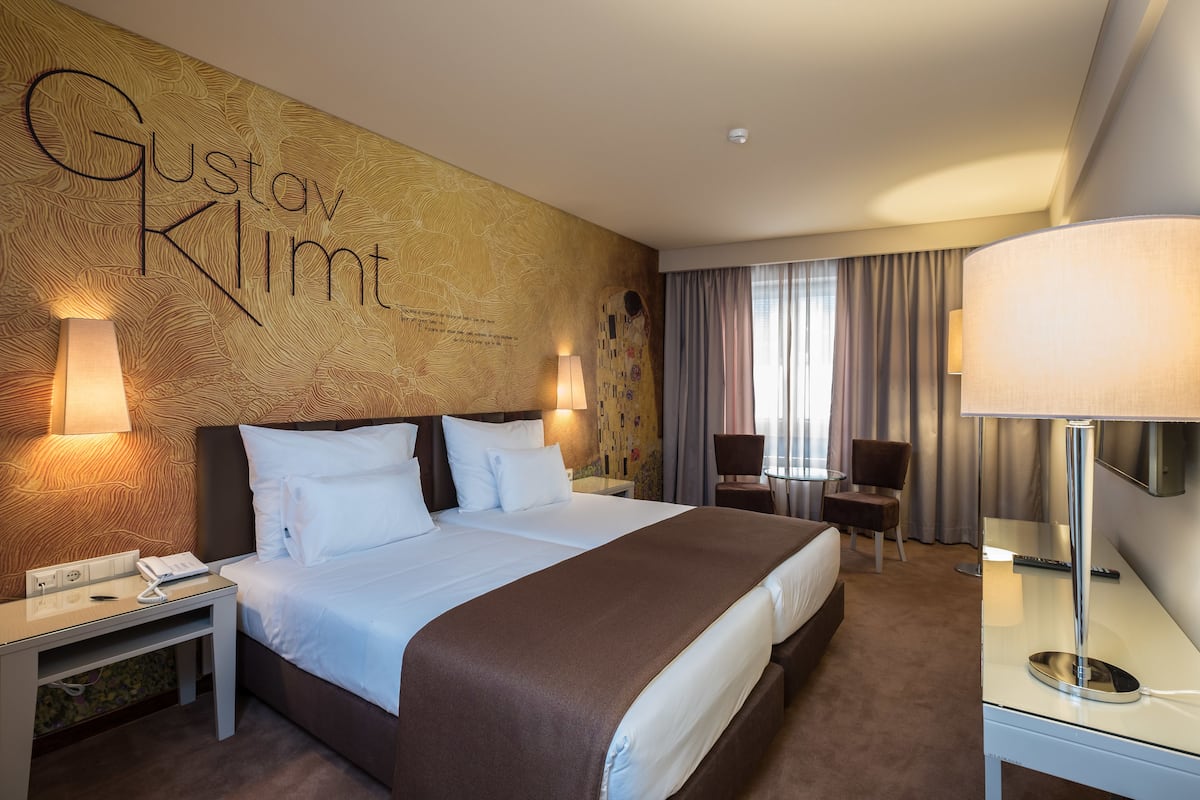 A hotel room with a large bed, two bedside tables, a desk, and a chair. The room has a patterned wall and a large window with curtains.