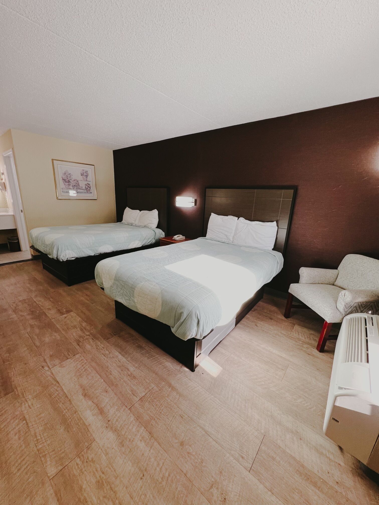 A hotel room with two beds, a chair, a bedside table with a phone, and a wall-mounted light.