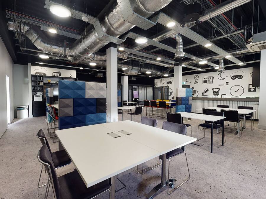 Un salón de conferencias moderno con mesas blancas y sillas negras, sistema de aire acondicionado visible y una pared con varios iconos y un reloj.