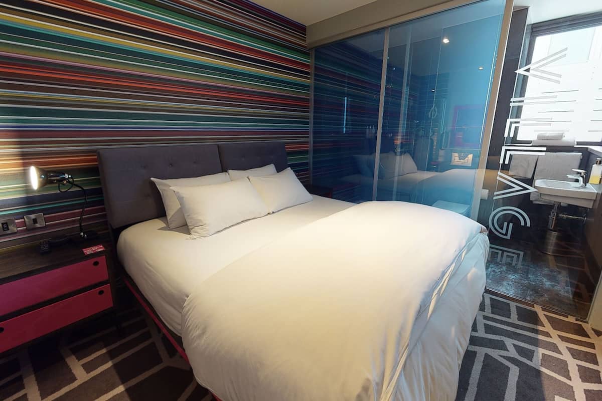 A modern hotel room with a large bed, a bedside table with a lamp, a glass partition, and a view of the bathroom.
