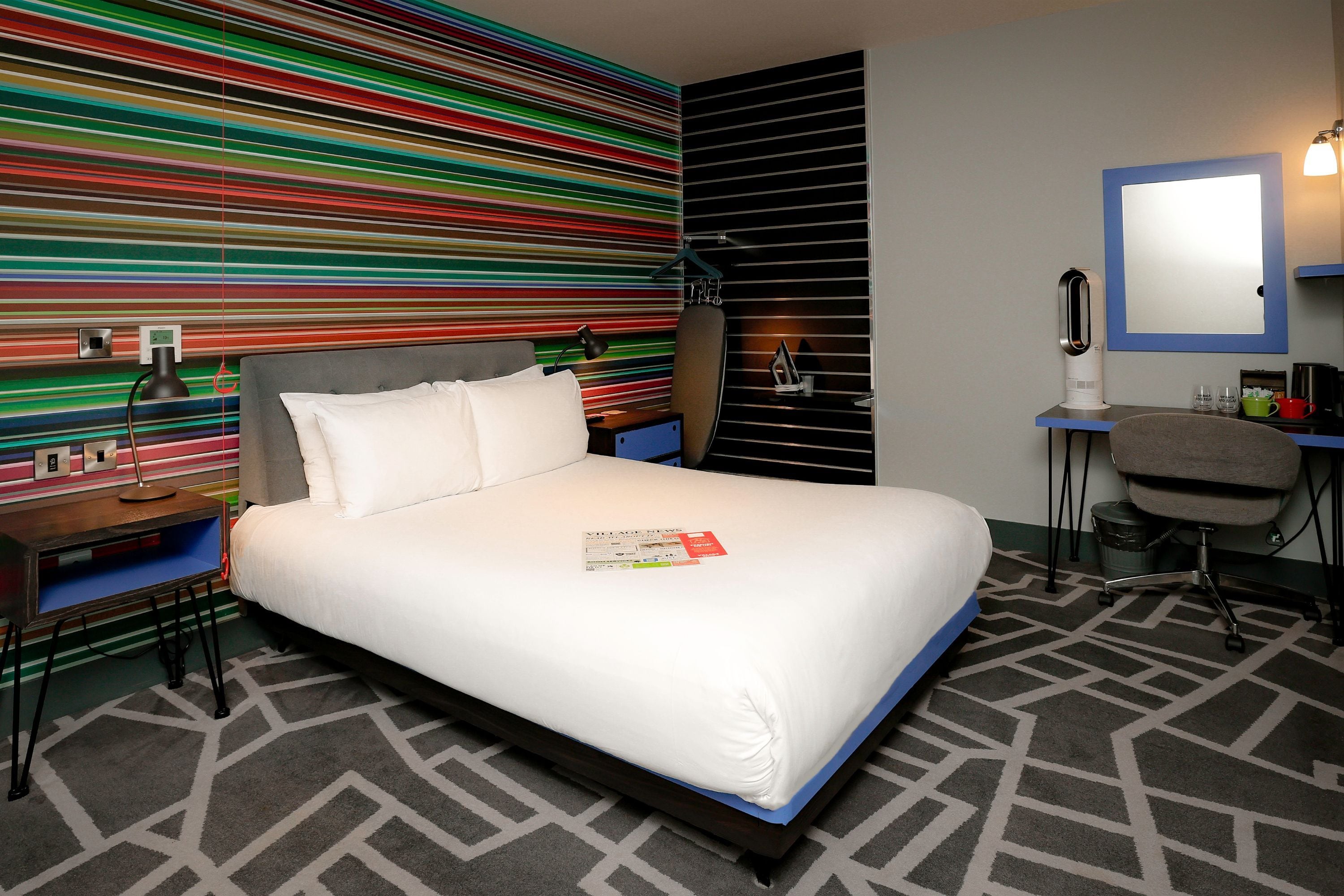 A hotel room with a bed, a desk, a chair, a mirror, and a colorful striped wall.