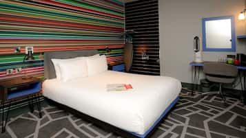 A hotel room with a bed, a desk, a chair, a mirror, and a colorful striped wall.
