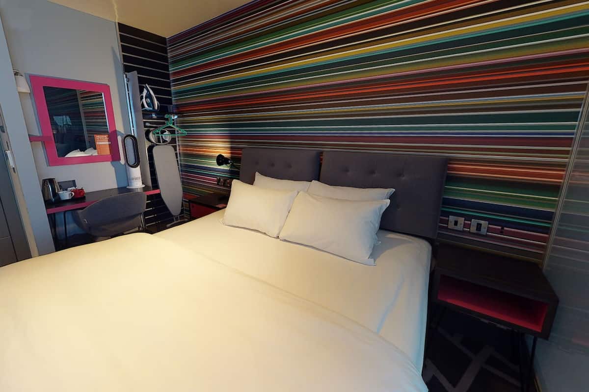 A modern bedroom with a large bed, a bedside table, a chair, a mirror, and a colorful striped wall.