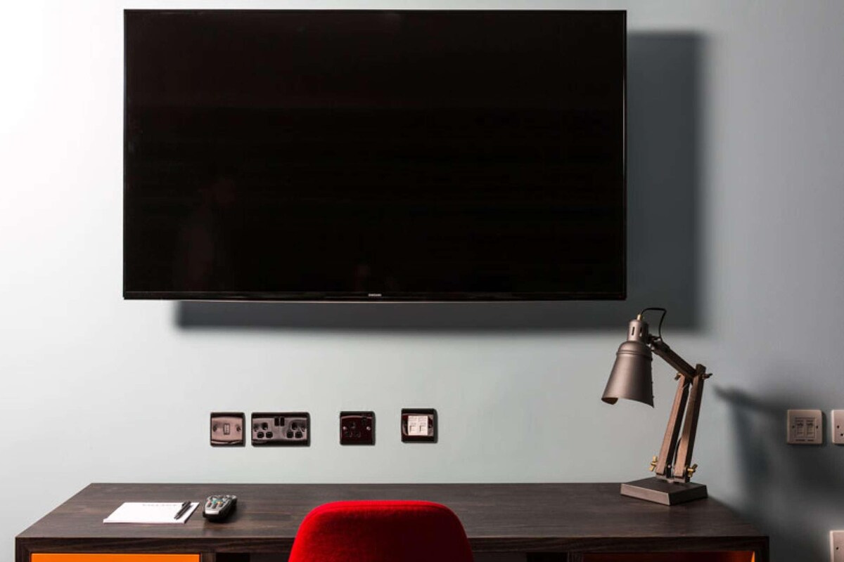Una habitación moderna con un televisor de pantalla plana montado en la pared, un escritorio con lámpara y una silla roja.
