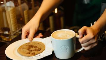 A person holding a coffee mug with latte art, a plate with a cookie, and a Starbucks cup.