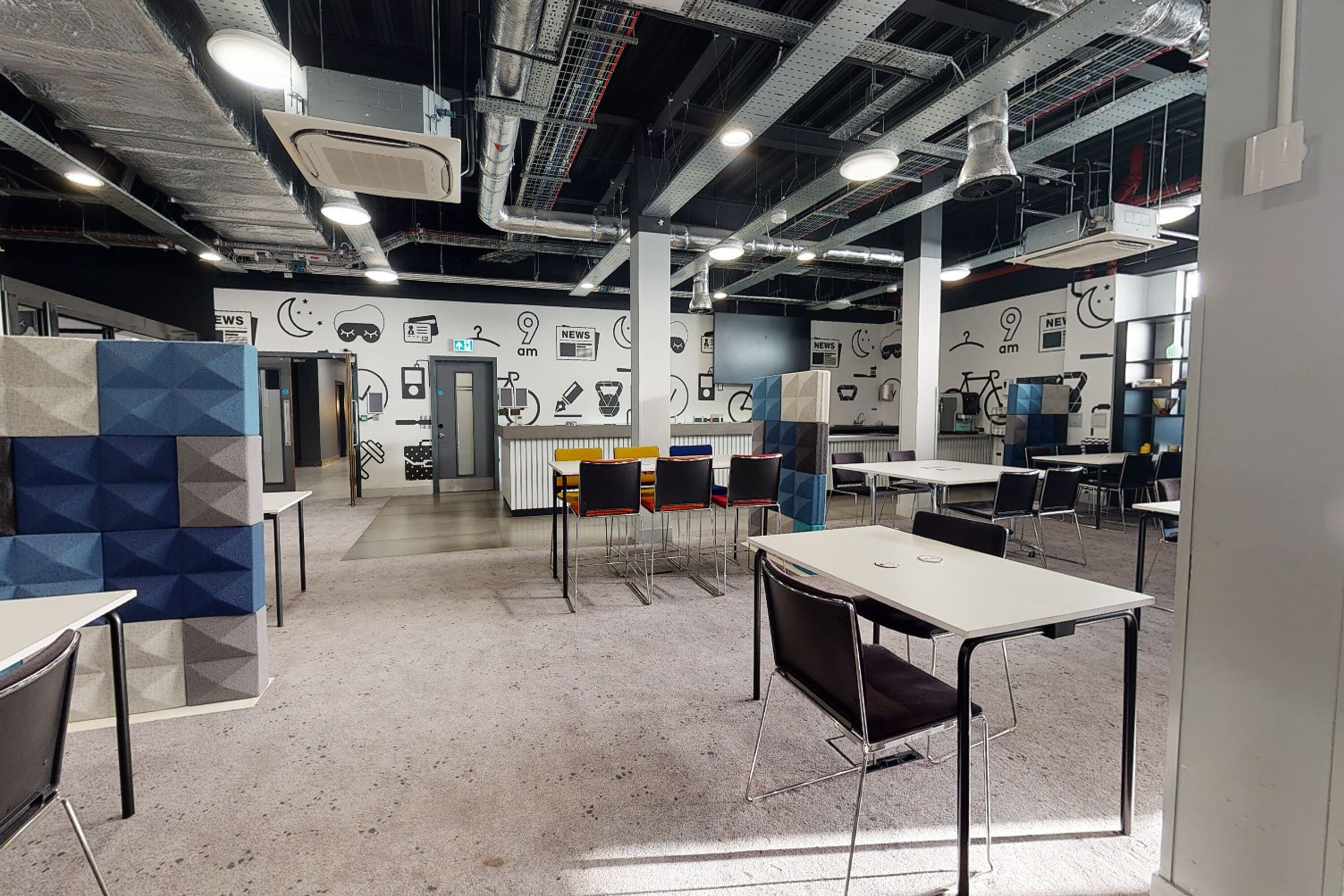 Un espacio de oficina moderno con varias mesas y sillas, un techo alto con conductos expuestos y una pared con un diseño que incluye varios iconos.