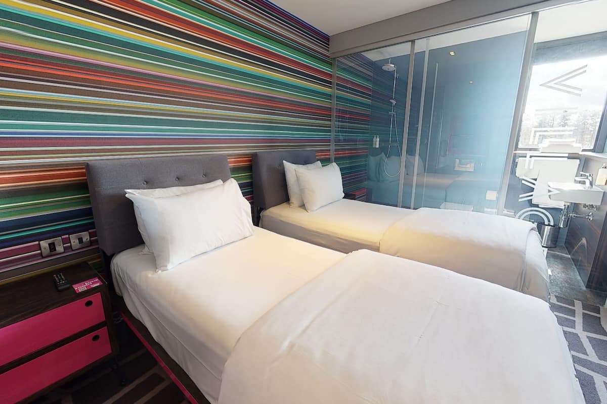 A hotel room with two beds, a colorful striped wall, a glass shower area, and a view of the outside.