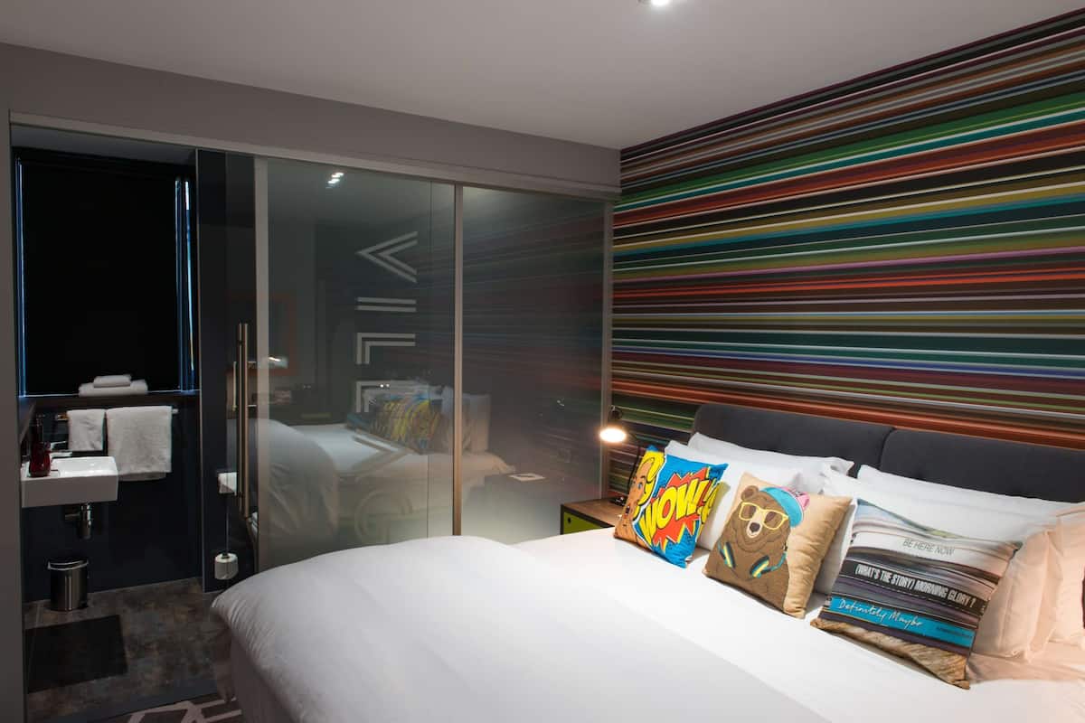 A modern hotel room with a large bed, a glass shower enclosure, and a colorful striped wall.