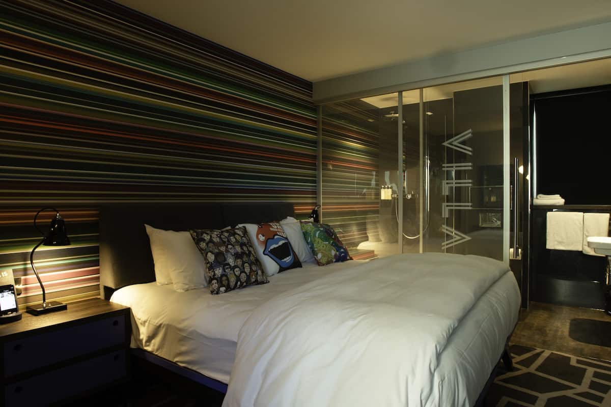 A modern hotel room with a large bed, a bedside table with a lamp, and a glass partitioned shower area.