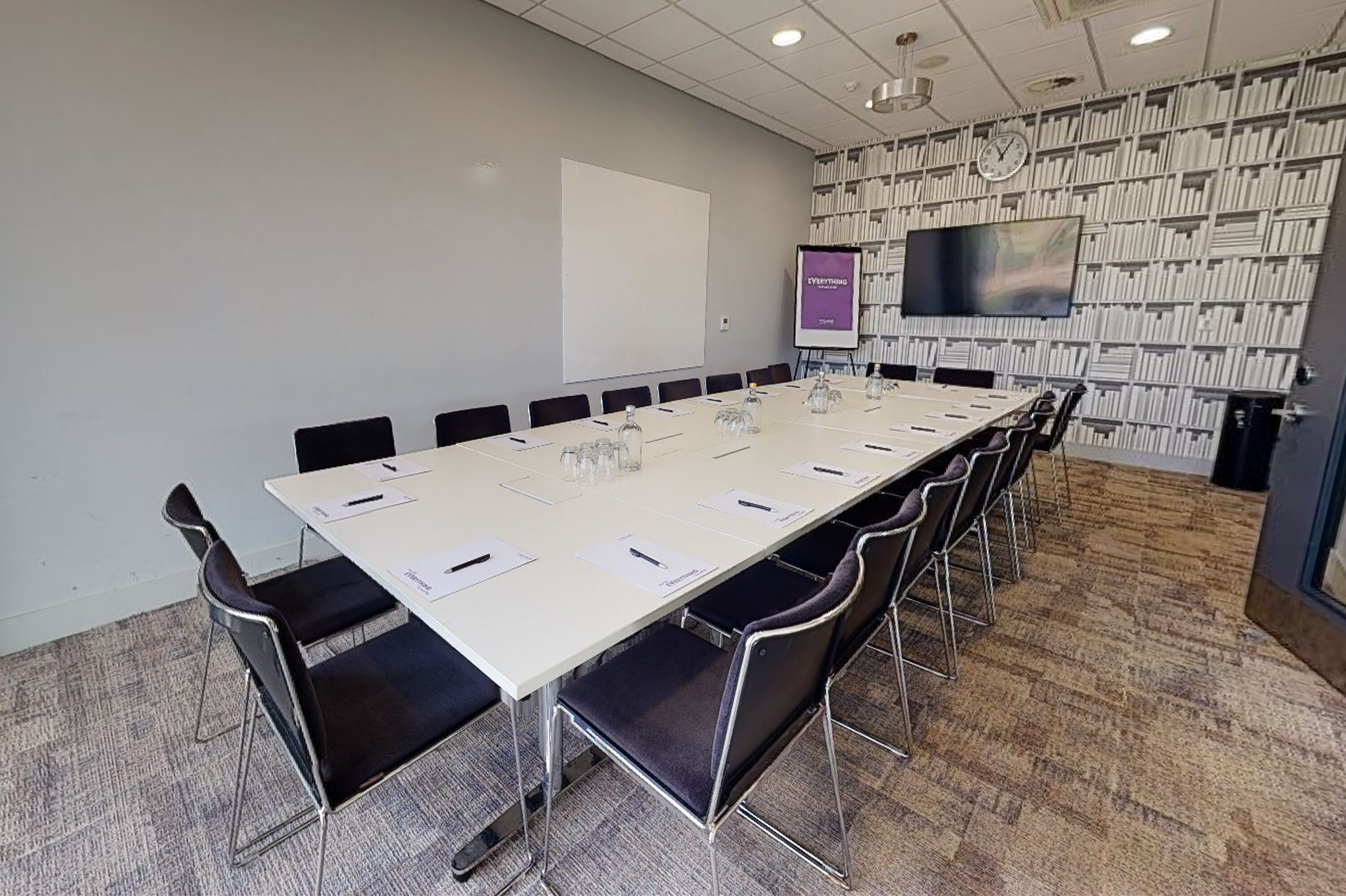Uma sala de conferências com uma mesa comprida, cadeiras, uma tela de projetor, um quadro branco e uma estante de livros.