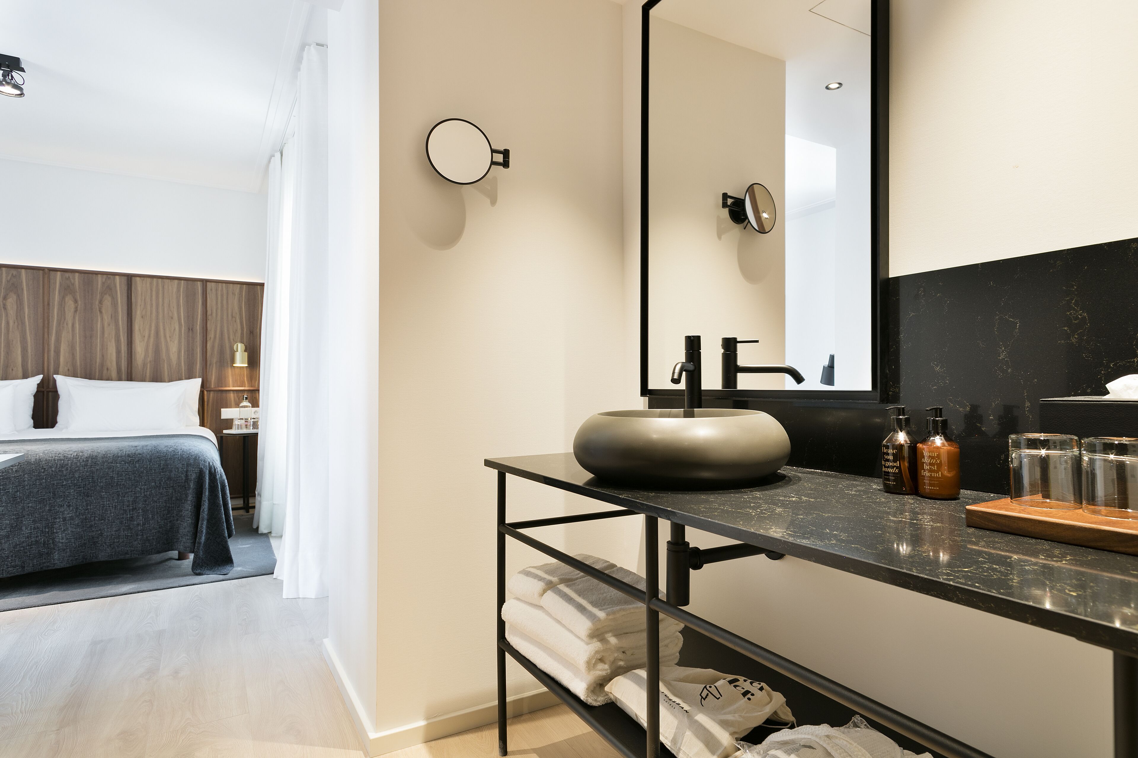A modern hotel room with a large bed, a round mirror, a black countertop with a sink, and neatly folded towels.