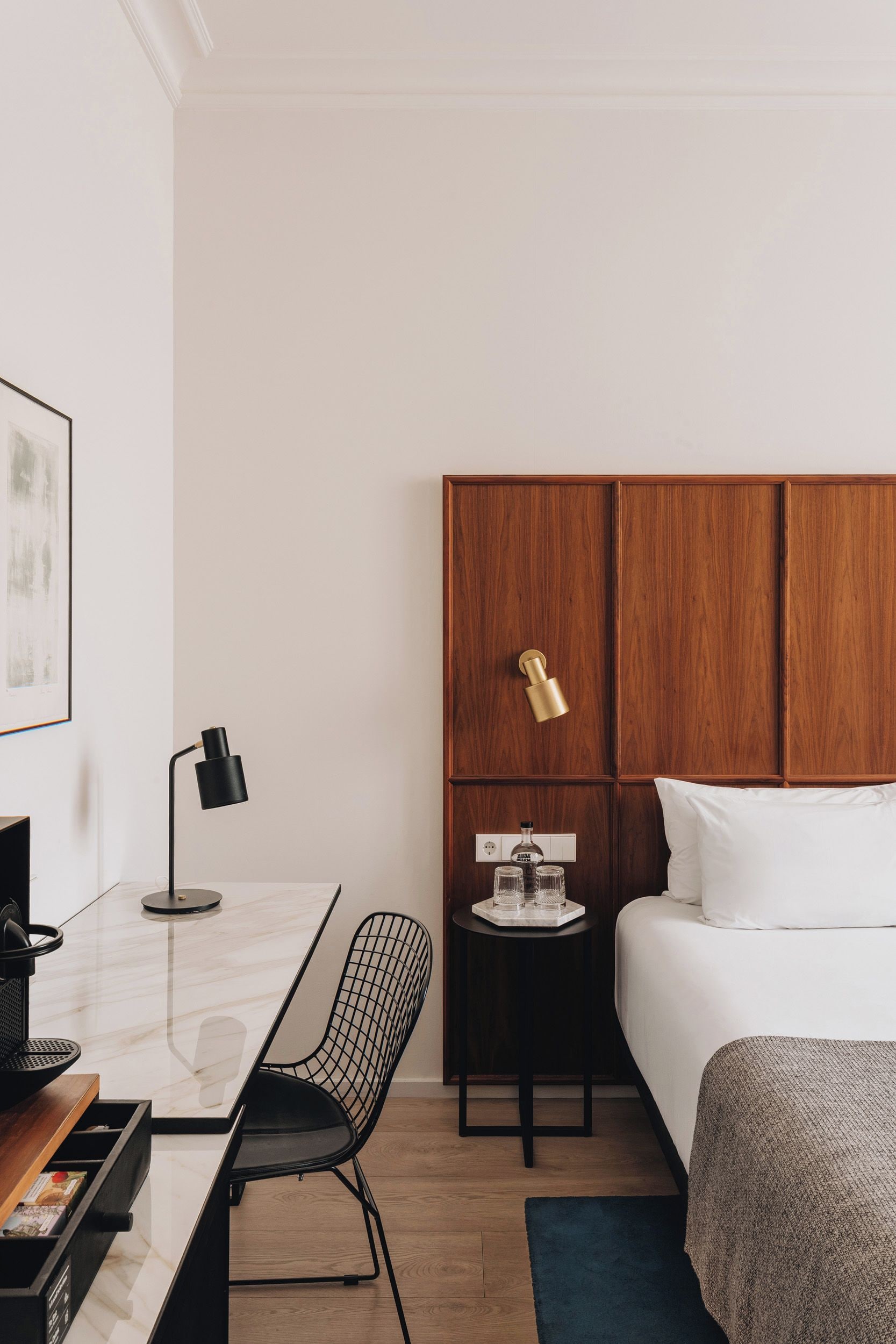 Habitación de hotel con cama, escritorio con silla, una mesita con vasos y una botella, y una lámpara de pared.