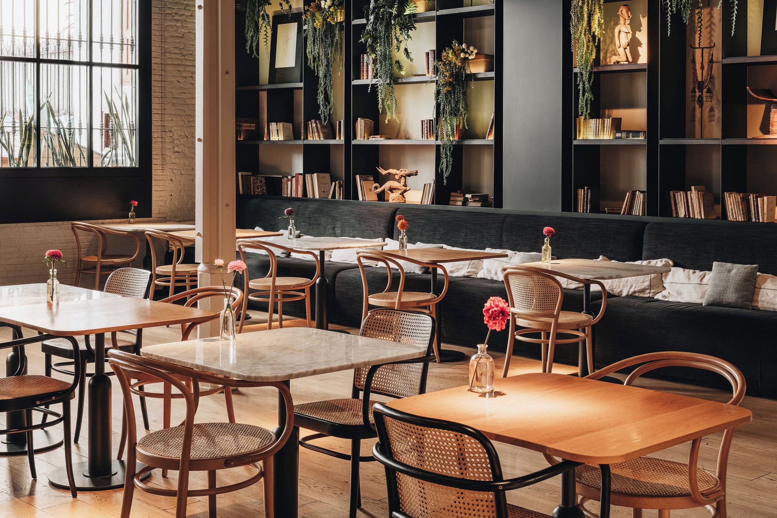 Un café chaleureux avec des tables et des chaises en bois, un canapé et des étagères remplies de plantes et de livres.