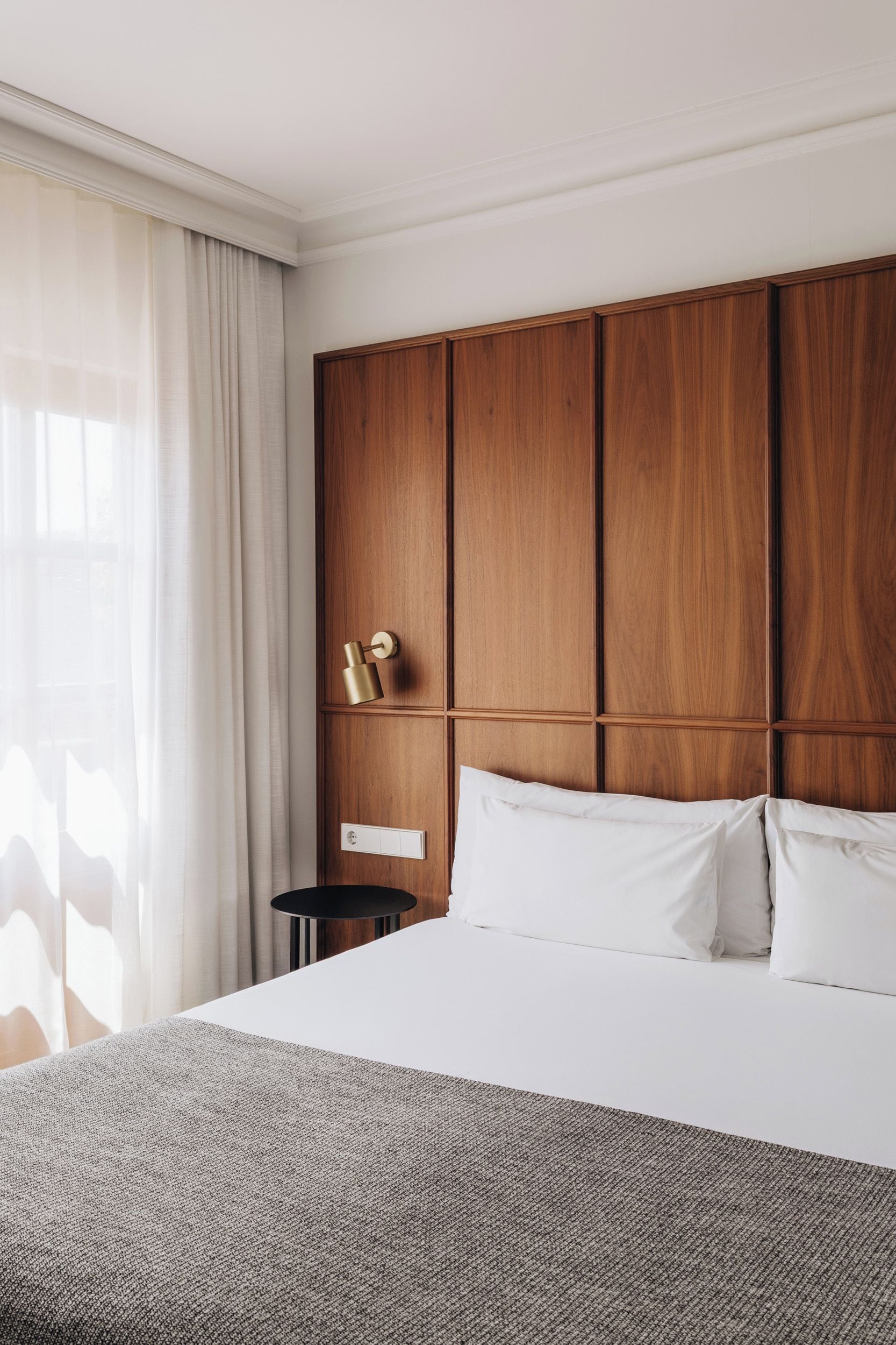 A hotel room with a large bed, wooden headboard, a bedside table, and a window with curtains.
