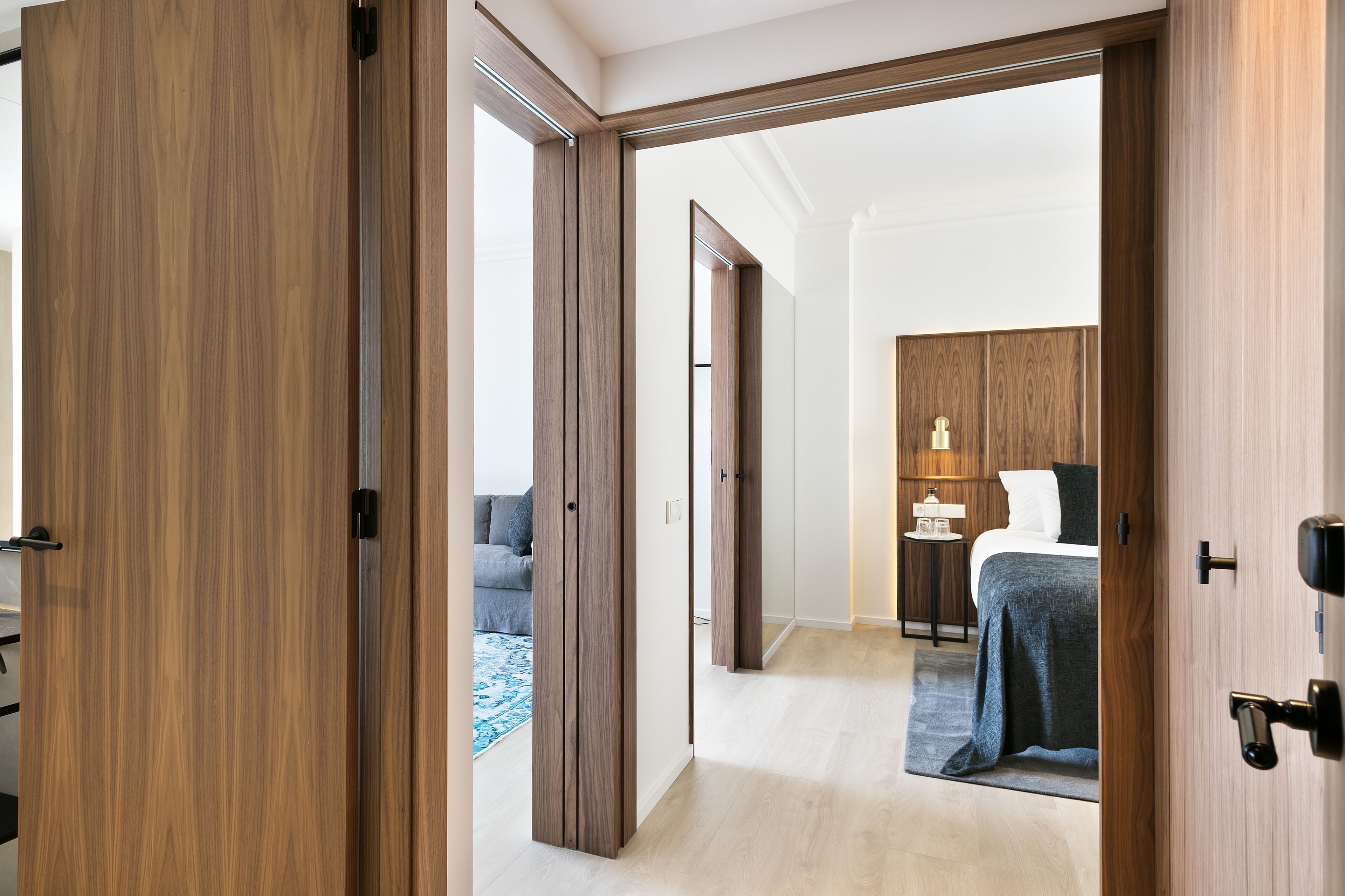 Une chambre d’hôtel avec une porte en bois, un lit recouvert d’une couverture grise, une table de chevet avec une lampe, et une vue sur une autre pièce où se trouvent un canapé et une table basse.