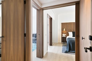 Hotelkamer met een houten deur, een bed met een grijze plaid, een nachtkastje met een lamp, en uitzicht op een andere kamer met een bank en een salontafel.