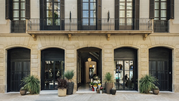 A building with three large arched doorways, a balcony with black railings, and potted plants in front.