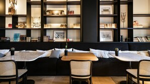 A modern interior with a black sofa, two tables, and wicker chairs. Shelves with books and decorative items are visible.