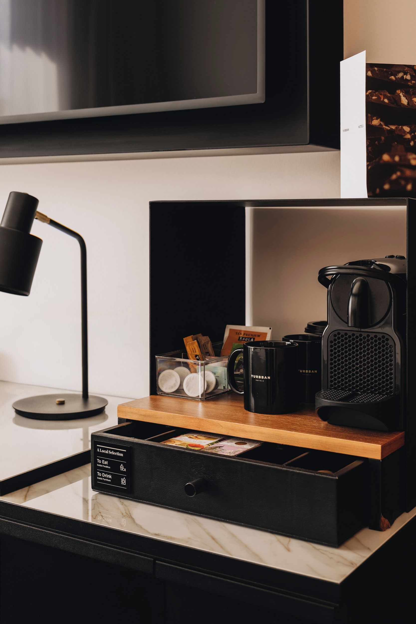 A modern hotel room with a flat-screen TV mounted on the wall, a black desk with a lamp, a coffee machine, and a drawer unit with a label indicating it contains food and drink items.