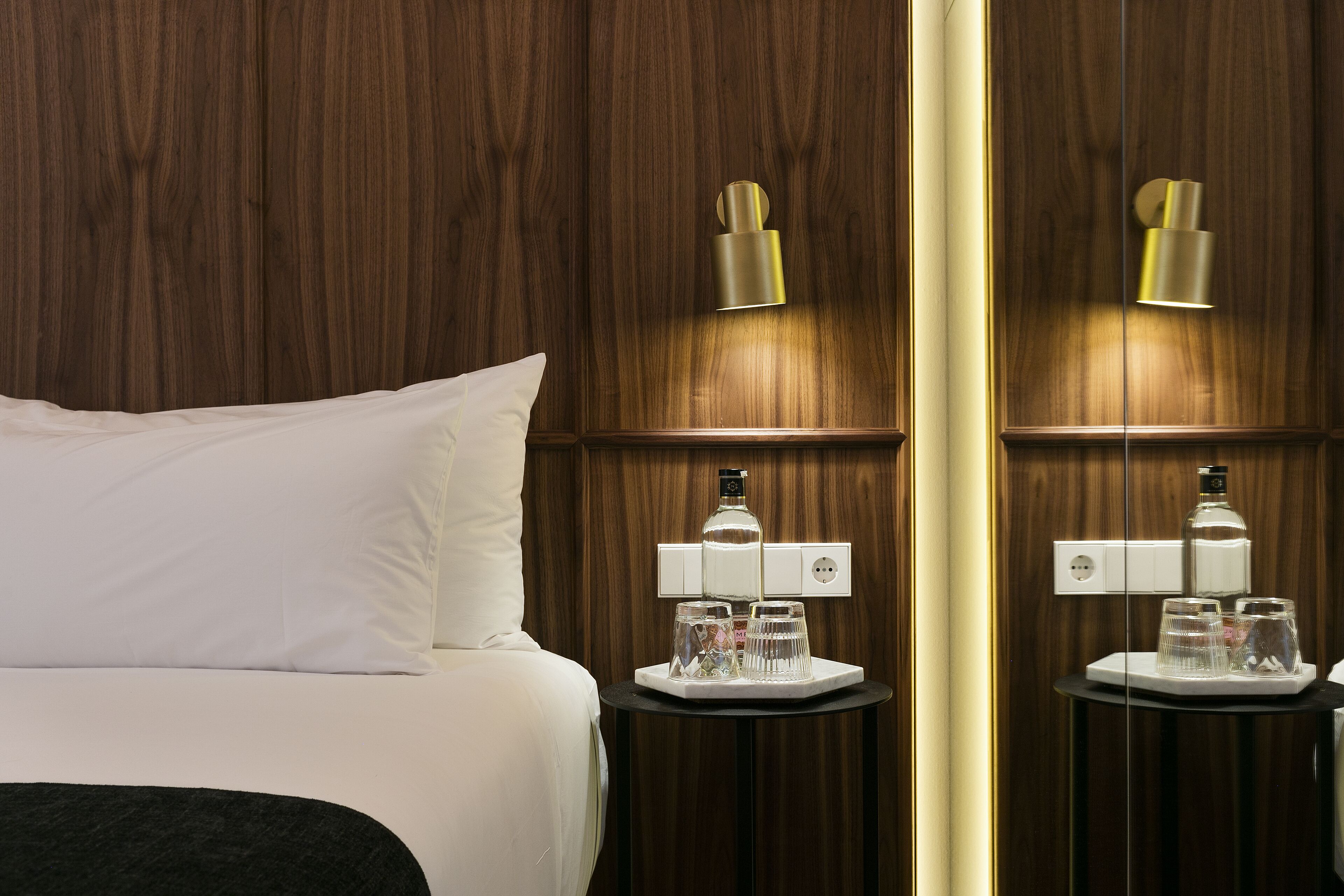 A hotel room with a bed, a bedside table with a lamp, and a mirror reflecting the room.
