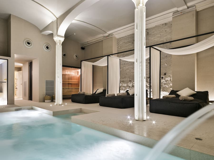 A modern indoor pool area with a jacuzzi, black sofas, and a large window with sheer curtains.