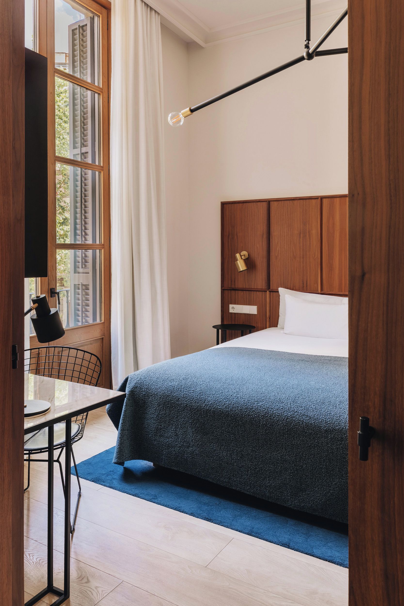 A hotel room with a bed, a desk, a chair, a window with curtains, and a lamp.