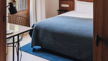 A hotel room with a bed, a desk, a chair, a window with curtains, and a lamp.