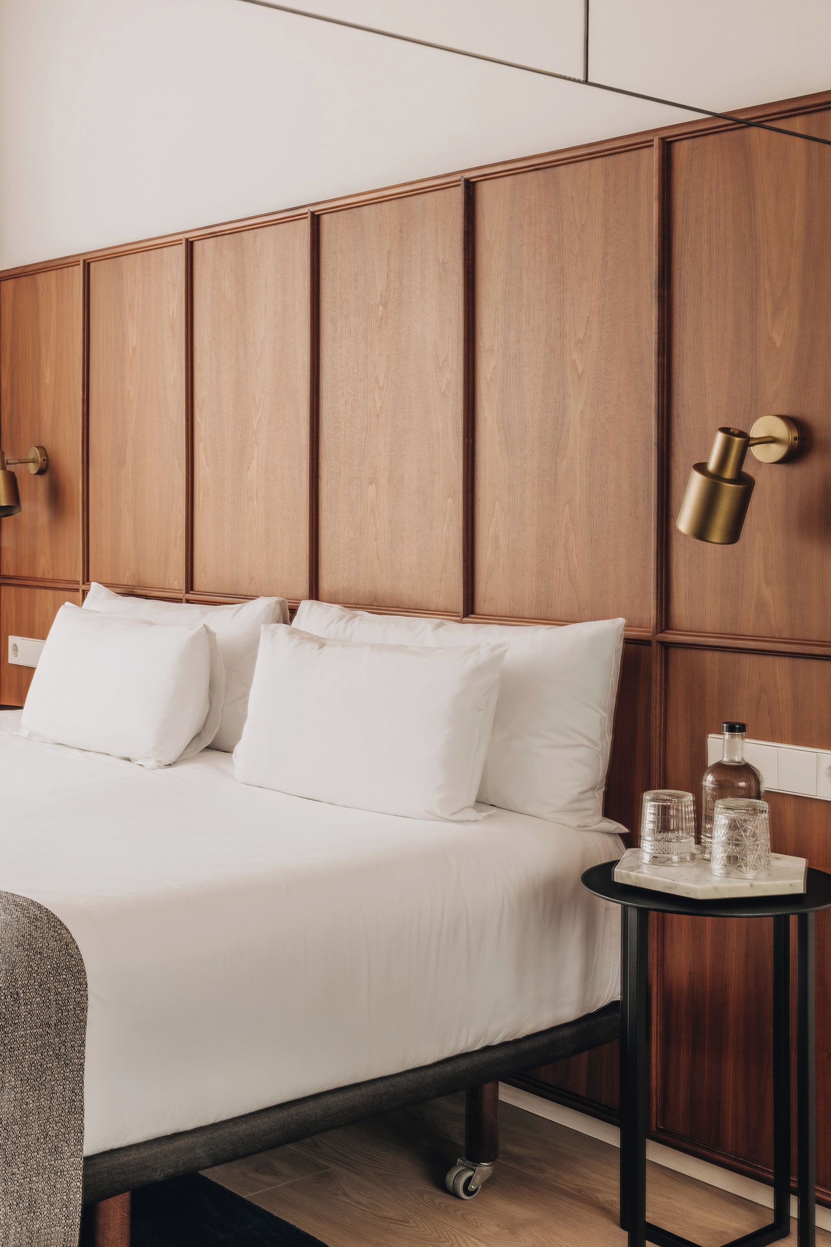 Habitación de hotel con cabecera de madera, cama con sábanas blancas, una mesita redonda con una botella y vasos, y una lámpara fijada a la pared.
