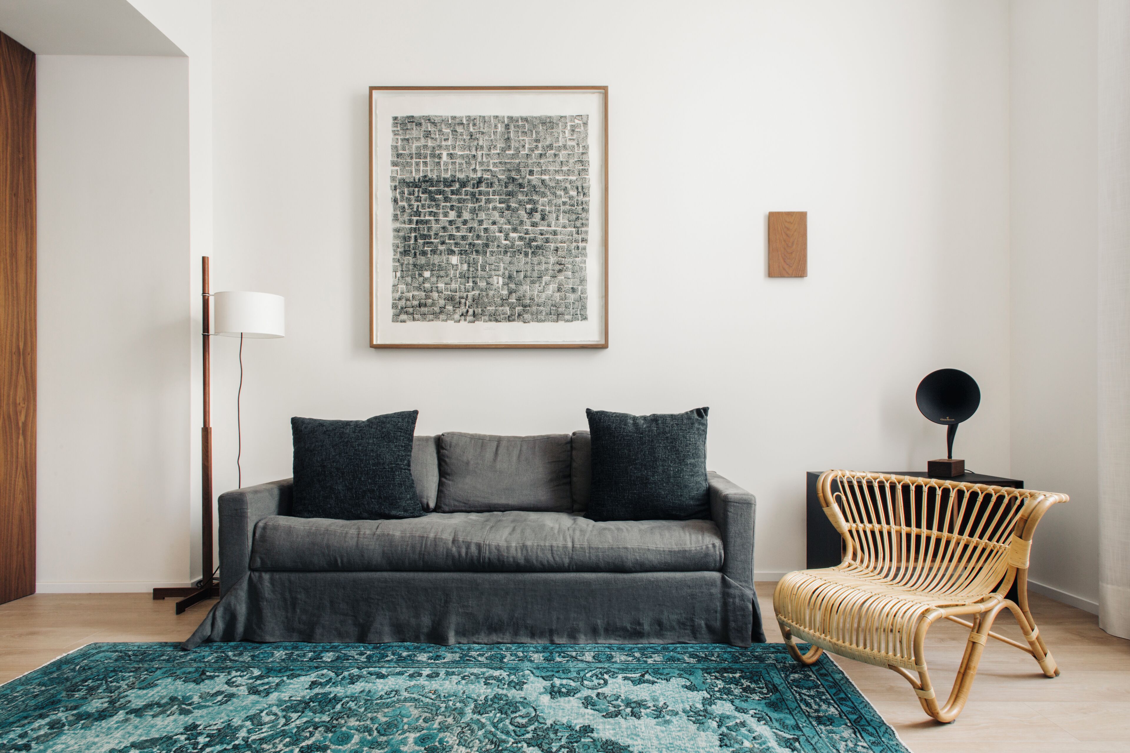 Ein Zimmer mit grauem Sofa, einem Korbstuhl, einem gemusterten Teppich und einem gerahmten abstrakten Kunstwerk an der Wand.